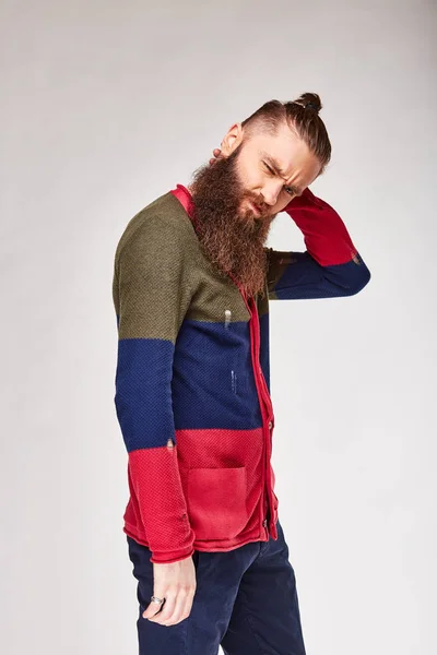 Молодой бородатый мужчина с модными волосами выглядит серьезным в студии в модном современном цветном свитере с красными, темно-синими и оливковыми полосками и синими чино — стоковое фото