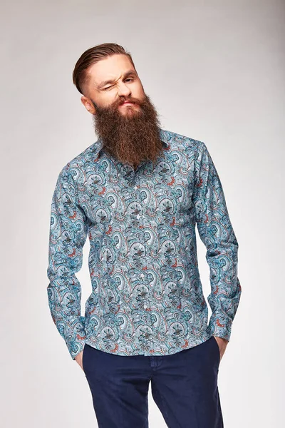 Joven hombre barbudo posando en estudio en camisa gris de moda con estampado y pantalones azules —  Fotos de Stock