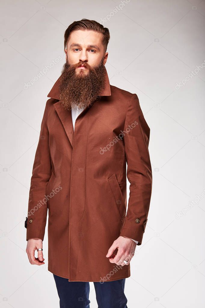 Young bearded man posing in studio in brown long jacket and blue pants