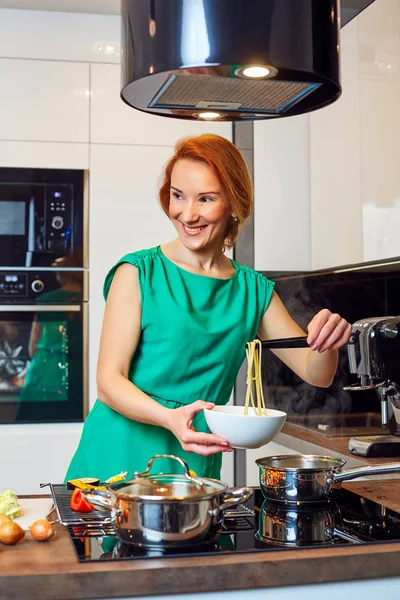 Pretty happy woman with red hair standing near table with different vegetables and holding bowl with pasta in high-tech modern kitchen