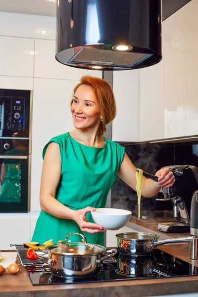 Pretty happy woman with red hair standing near table with different vegetables and holding bowl with pasta in high-tech modern kitchen
