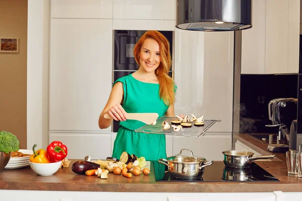 Pretty smiling woman with red hair standing near kitchen table and holding grille with vegetables, looking at the camera in high-tech modern sunny kitchen