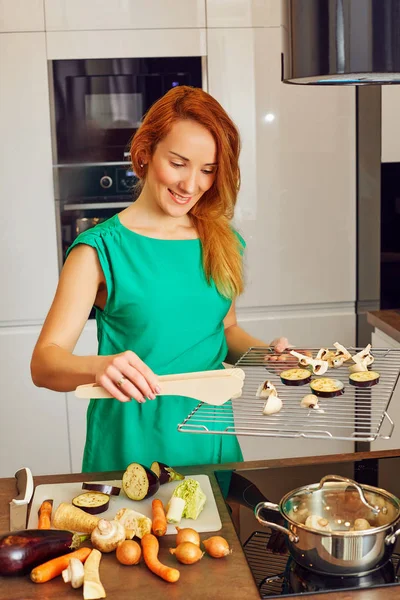 Pretty smiling woman with red hair standing near kitchen table and holding grille with vegetables, cooking in high-tech modern sunny kitchen