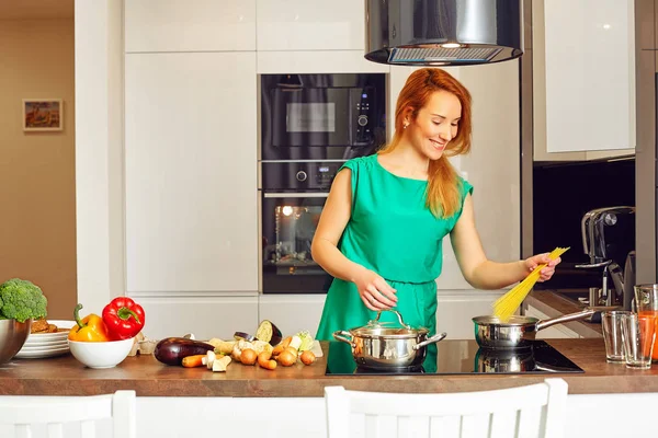 Happy smiling attentive woman with red hair standing near kitchen table with colorful vegetables and puts pasta into the pan, cooking in high-tech modern sunny kitchen