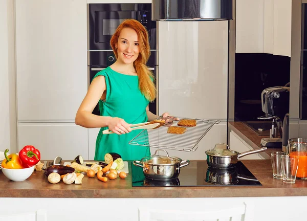 Happy smiling attentive woman with red hair standing near kitchen table with colorful vegetables and puts toasts on the grille, cooking in high-tech modern sunny kitchen and looking at camera