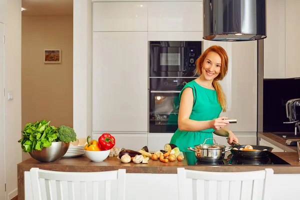 Happy smiling attentive woman with red hair standing near kitchen table with colorful vegetables and cooking in high-tech modern sunny kitchen