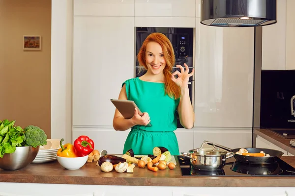 Happy smiling attentive woman with red hair standing near kitchen table with colorful vegetables and cooking in high-tech modern sunny kitchen, showing all right