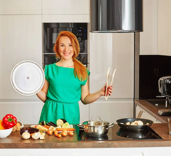 Happy smiling attentive woman with red hair standing near kitchen table with colorful vegetables with cap pot and tongs in high-tech modern sunny kitchen