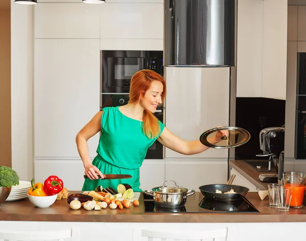 Pretty smiling attentive woman with red hair standing near kitchen table with colorful vegetables with cap pot and cooking in high-tech modern sunny kitchen