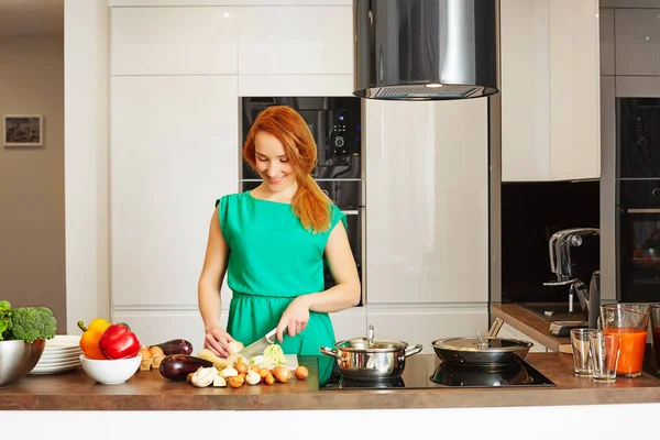 Happy smiling attentive woman with red hair standing near kitchen table with colorful vegetables and cutting vegetables, cooking in high-tech modern sunny kitchen