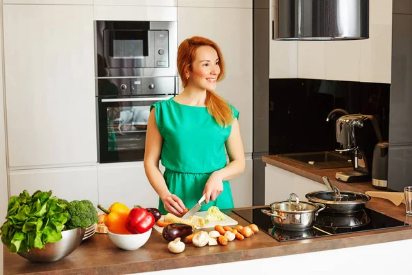 Happy smiling woman with red hair standing near kitchen table with colorful vegetables and cutting vegetables, cooking in high-tech modern sunny kitchen and looking away