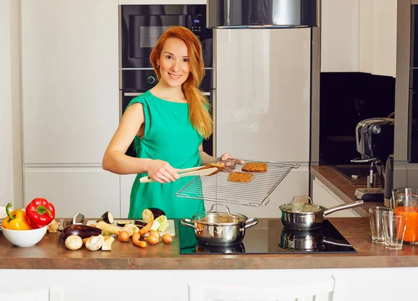 Happy smiling attentive woman with red hair standing near kitchen table with colorful vegetables and puts toasts on the grille, cooking in high-tech modern sunny kitchen and looking at camera Stock Picture