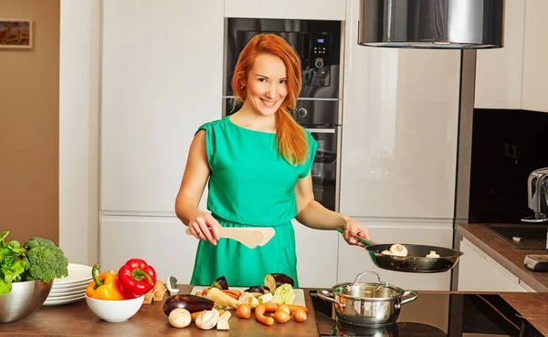 Wanita yang tersenyum penuh perhatian dengan rambut merah berdiri di dekat meja dapur dengan sayuran berwarna-warni dengan penggorengan yang diisi dengan jamur dan memasak di dapur modern berteknologi tinggi yang cerah Stok Foto