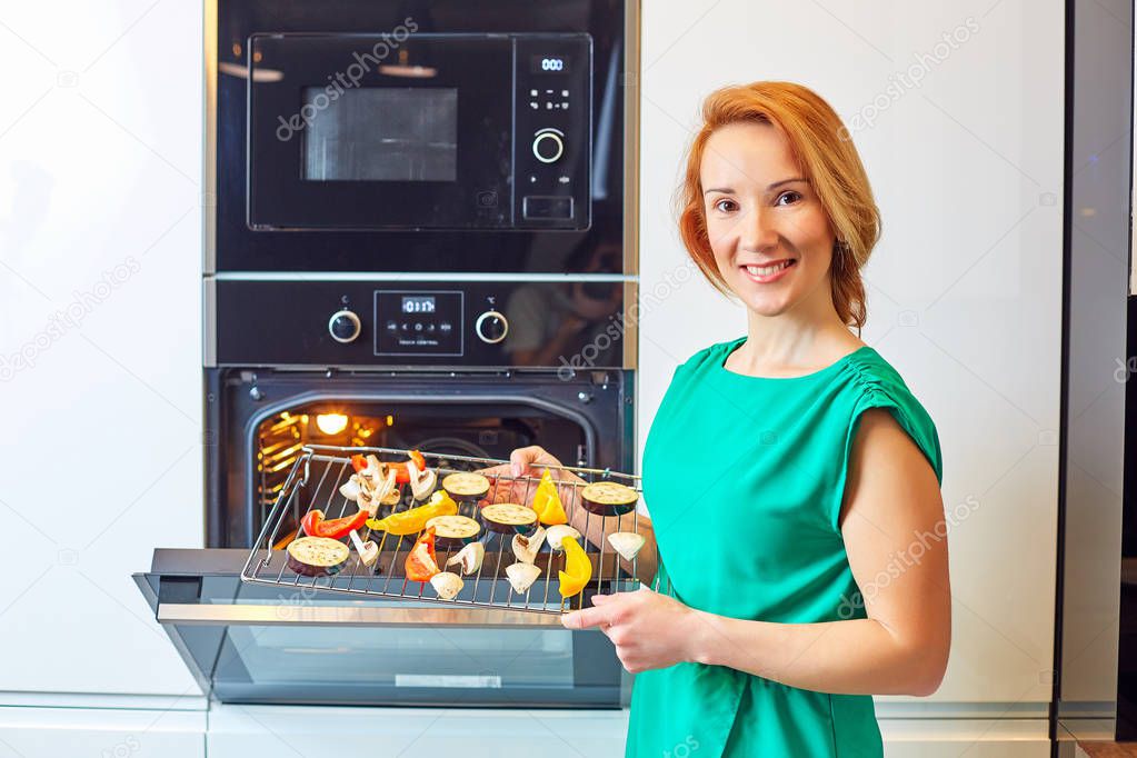 Young smiling woman with red hair standing near oven and holding grille with vegetables, looking at camera in modern kitchen