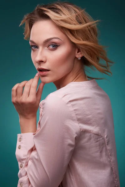 Potret wanita muda berambut pirang yang romantis dengan mata besar dan rambut bergelombang, mengenakan gaun merah muda terang di studio dengan latar belakang pirus yang mendalam — Stok Foto