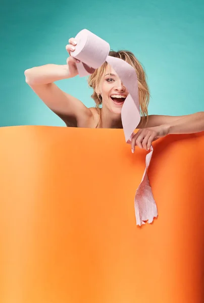 Joven mujer rubia sonriente jugando con rollo de papel higiénico en el estudio sobre fondo turquesa y hoja naranja —  Fotos de Stock