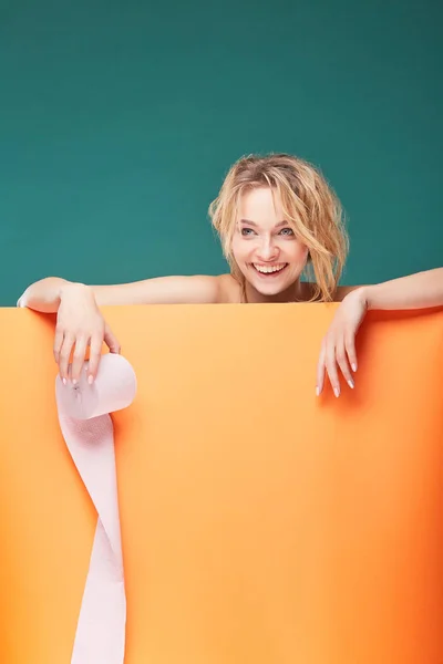 Joven hermosa mujer rubia sosteniendo rollo de papel higiénico con la cara sonriente en el estudio sobre fondo turquesa y hoja naranja —  Fotos de Stock