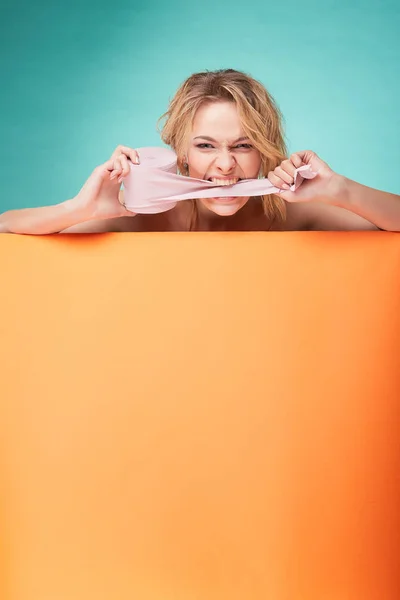 Joven hermosa mujer rubia sosteniendo rollo de papel higiénico con la cara de risa y mordiéndolo en el estudio sobre fondo turquesa y hoja naranja —  Fotos de Stock