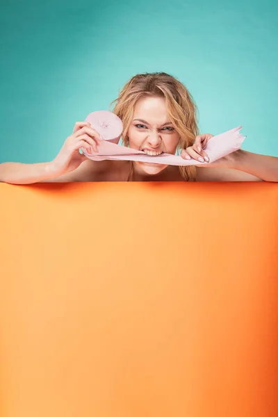 Joven hermosa mujer rubia sosteniendo rollo de papel higiénico con la cara de risa y mordiéndolo en el estudio sobre fondo turquesa y hoja naranja —  Fotos de Stock