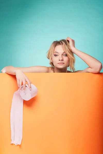 Joven hermosa mujer rubia sosteniendo rollo de papel higiénico con cara de pensamiento en el estudio sobre fondo turquesa y hoja naranja —  Fotos de Stock