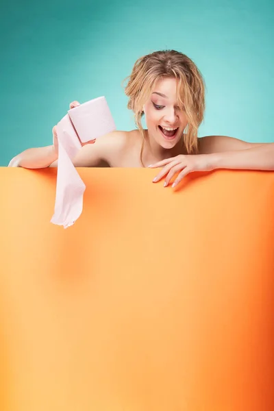 Νέοι χαριτωμένο ξανθιά γυναίκα κρατώντας ρολό χαρτί τουαλέτας και κοιτάζοντας προς τα κάτω με χαμογελαστό πρόσωπο στο studio σε φόντο τιρκουάζ και πορτοκαλί φύλλο — Φωτογραφία Αρχείου