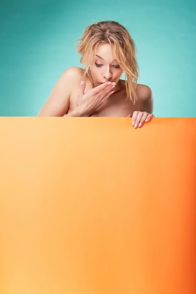 Young surprised blonde woman posing in studio with turquoise background and orange sheet — Stock Photo, Image