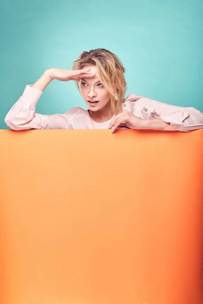 Mujer rubia linda joven buscando algo en el estudio con fondo turquesa y hoja naranja —  Fotos de Stock