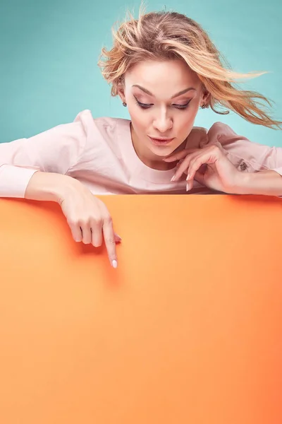 Retrato de una joven rubia increíble mirando hacia abajo y mostrando algo en el estudio con fondo turquesa y sábana naranja —  Fotos de Stock