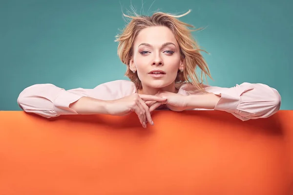 Portrait of young amazing blonde woman in light pink dress looking at camera in studio with turquoise background and orange sheet — Stock Photo, Image