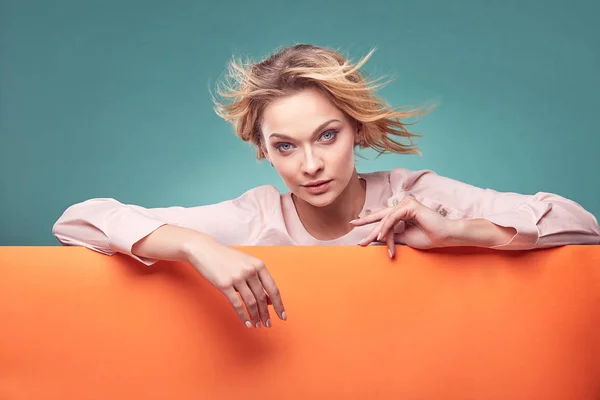 Portrait of young sexy blonde woman in light pink dress looking at camera in studio with turquoise background and orange sheet — Stock Photo, Image