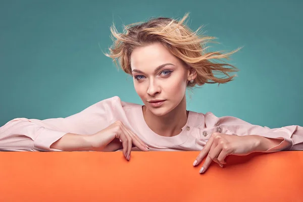 Portrait of young pretty blonde woman in light pink dress looking with interest in studio with turquoise background and orange sheet — Stock Photo, Image