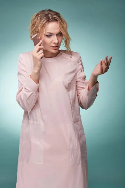 光条粉红色的裙子用粉色智能手机通话和看上去忙碌演播室青绿色背景上的波浪式头发的年轻漂亮金发女子 — 图库照片