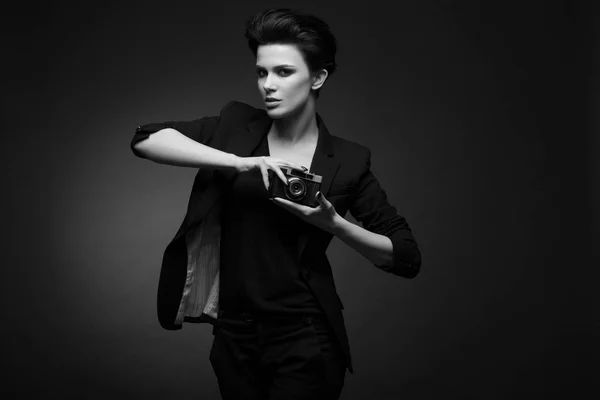 Wanita cantik serius dengan rambut hitam pendek mengenakan blazer hitam berpose di studio gelap, memegang kamera foto tua di tangannya, hitam dan putih — Stok Foto