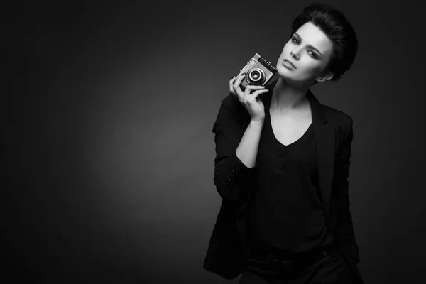 Молодая серьезная красивая женщина с короткими темными волосами в черном ретро-блейзере позирует в темной студии, держа в руках старую фотокамеру, в черно-белом — стоковое фото