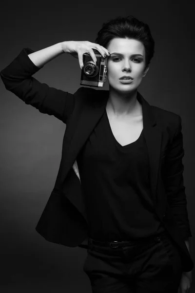 Молодая серьезная удивительная женщина с короткими темными волосами в черном ретро-блейзере позирует в темной студии, держа в руках старую фотокамеру, в черно-белом — стоковое фото