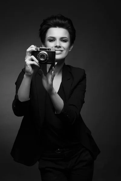 Young sexy woman photographer with short dark hair wearing black retro blazer posing in dark studio, holding old photo camera in her hands, in black and white — Stock Photo, Image