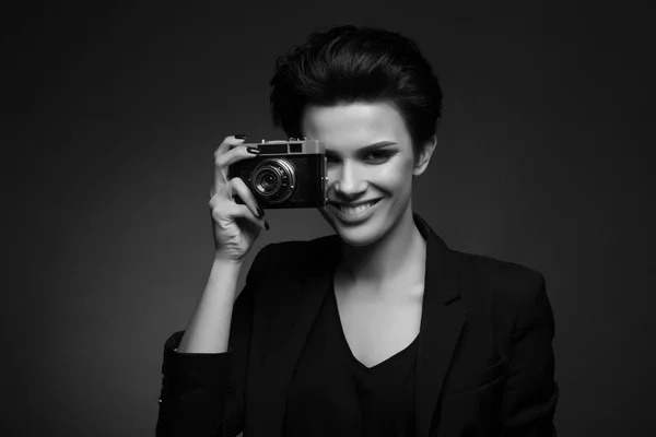 Jeune photographe souriante aux cheveux bruns courts portant un blazer rétro noir posant en studio sombre, montrant un vieil appareil photo dans ses mains, en noir et blanc — Photo