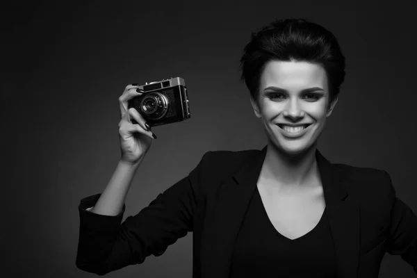 Jeune photographe souriante aux cheveux bruns courts portant un blazer rétro noir posant en studio sombre, montrant un vieil appareil photo dans ses mains, en noir et blanc — Photo