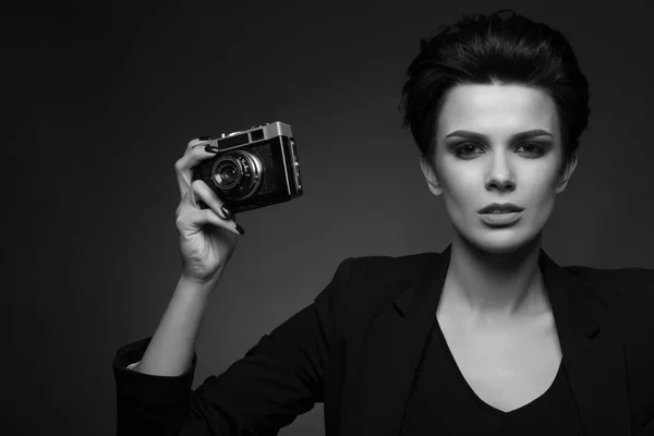 Tânără femeie drăguță fotograf cu păr scurt închis la culoare purtând blazer retro negru pozând în studio întunecat, arătând camera foto veche în mâinile ei, în alb și negru — Fotografie, imagine de stoc