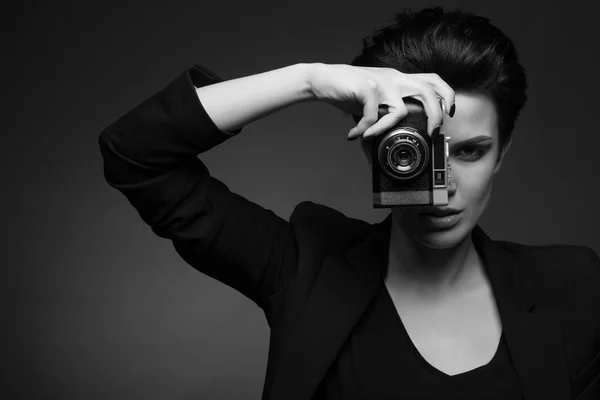 若いかわいい女性カメラマン短い黒い髪を着て黒レトロなブレザー黒と白で、彼女の手で古い写真をカメラで暗いスタジオでポーズ — ストック写真