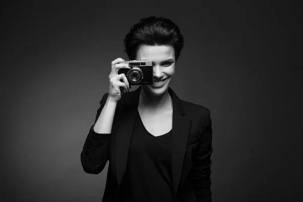 Młodych cute uśmiechający się kobieta fotograf na sobie krótkie włosy ciemny czarny retro blazer pozowanie Studio ciemny z stary aparat fotograficzny w jej ręce, czarno-białe — Zdjęcie stockowe