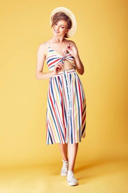 Kısa saçlı renkli yazlık elbise, hasır şapka ve Sarı zemin üzerine Studio'da poz beyaz ayakkabı giyen genç gülümseyen güzel kadın