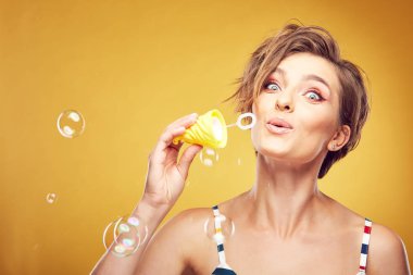 Seksi genç güzel kadın modeli ile kısa portresi closeup yüzü gülen ve kabarcıklar, Studio Sarı zemin üzerine poz üfleme ile hayran Kahverengi saç seyir darmadağınık