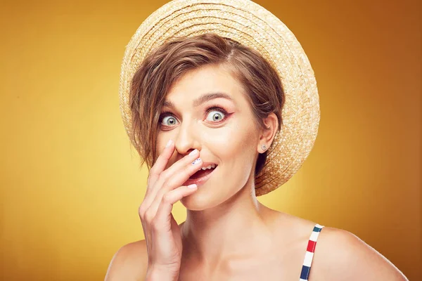 Retrato de cerca de la mujer bonita joven sexy con el pelo castaño desaliñado corto, mirando sorprendido con la mano cerca de la boca, usando sombrero de paja, posando en el estudio sobre fondo amarillo —  Fotos de Stock