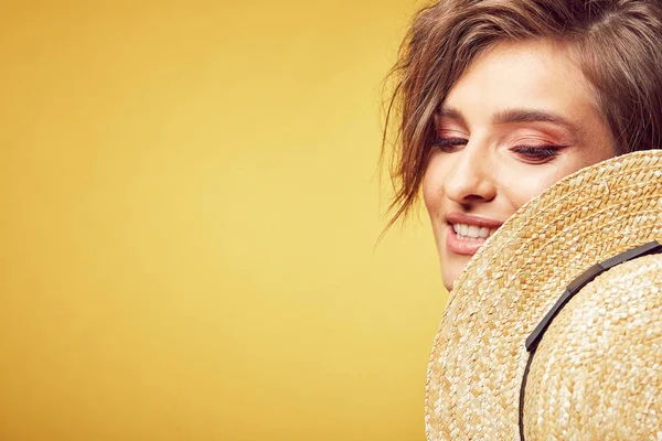 Closeup portrait of sexy young pretty woman with short disheveled brown hair looking down with happy face, holding straw hat, posing in studio on yellow background — Stock Photo, Image