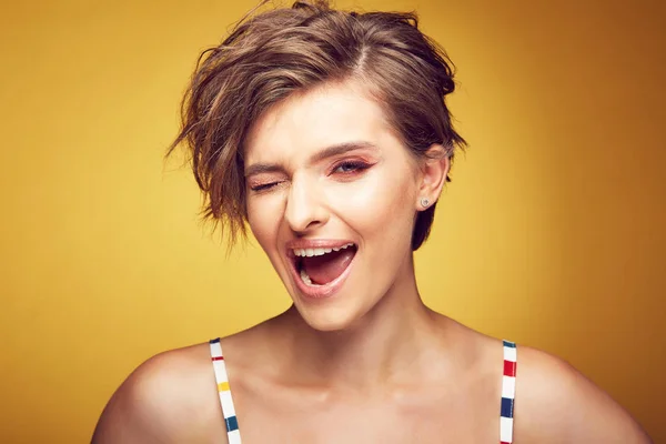 Primer plano retrato de sexy feliz joven modelo bastante femenina con pelo castaño desaliñado corto, y la cara sonriente, un ojo está cerrado, posando en el estudio sobre fondo amarillo — Foto de Stock