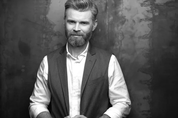 Amazing portrait of handsome charismatic serious man with grey beard, wearing white shirt and black waistcoat, posing  in studio on dark grey background in black and white — Stock Photo, Image