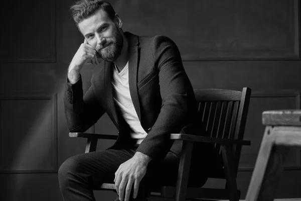 Bello giovane uomo barbuto bello e sicuro di sé, con camicia bianca e giacca marrone, seduto su una sedia di legno in studio con sfondo scuro e sorridente, in bianco e nero — Foto Stock