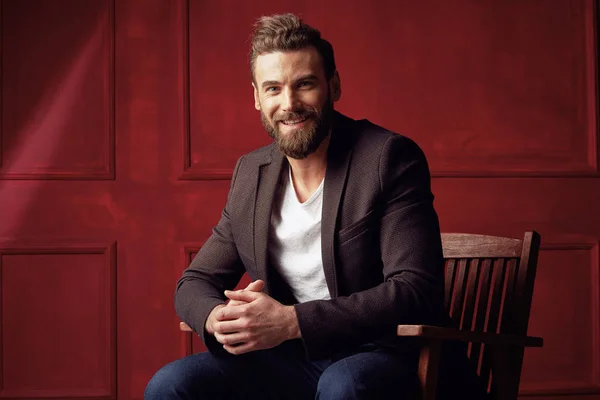Красивий бородатий усміхнений чоловік, одягнений у білу сорочку і коричневий піджак, сидить на дерев'яному стільці в темно-червоній студії — стокове фото