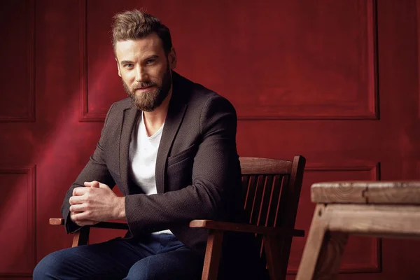Bonito homem barbudo bonito, vestindo camisa branca e casaco marrom, sentado em cadeira de madeira em estúdio com fundo vermelho escuro — Fotografia de Stock